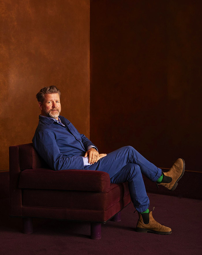 This image shows Tim Ross with short hair and a beard, seated on a plush, dark red armchair against a rich brown background. He is dressed in matching blue jacket and pants, paired with tan suede boots and bright green socks. He appears relaxed, leaning back slightly with his legs crossed, looking directly at the camera. The lighting highlights the textures of both the wall and his clothing with a warm tone.