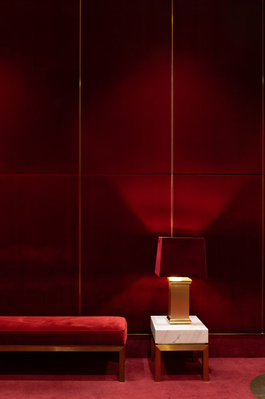 This image features an elegant and minimalistic interior design within the Arts Centre Melbourne Theatres building with a deep red color theme. A red velvet bench is positioned on the left side, and next to it, a modern table lamp with a rectangular gold base and a red lampshade sits atop a white marble side table. The background wall is paneled with smooth red velvet, and the overall lighting creates a warm, luxurious ambiance.